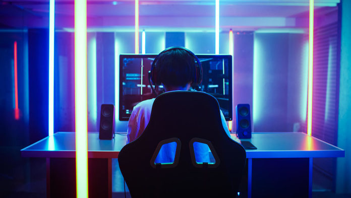 Back View Shot of the Beautiful Professional Gamer Girl Putting on Headset and Starts Playing Online Video Game on Her Personal Computer. Cute Casual Geek Girl. Room Lit by Neon Lamps in Retro Arcade Style.